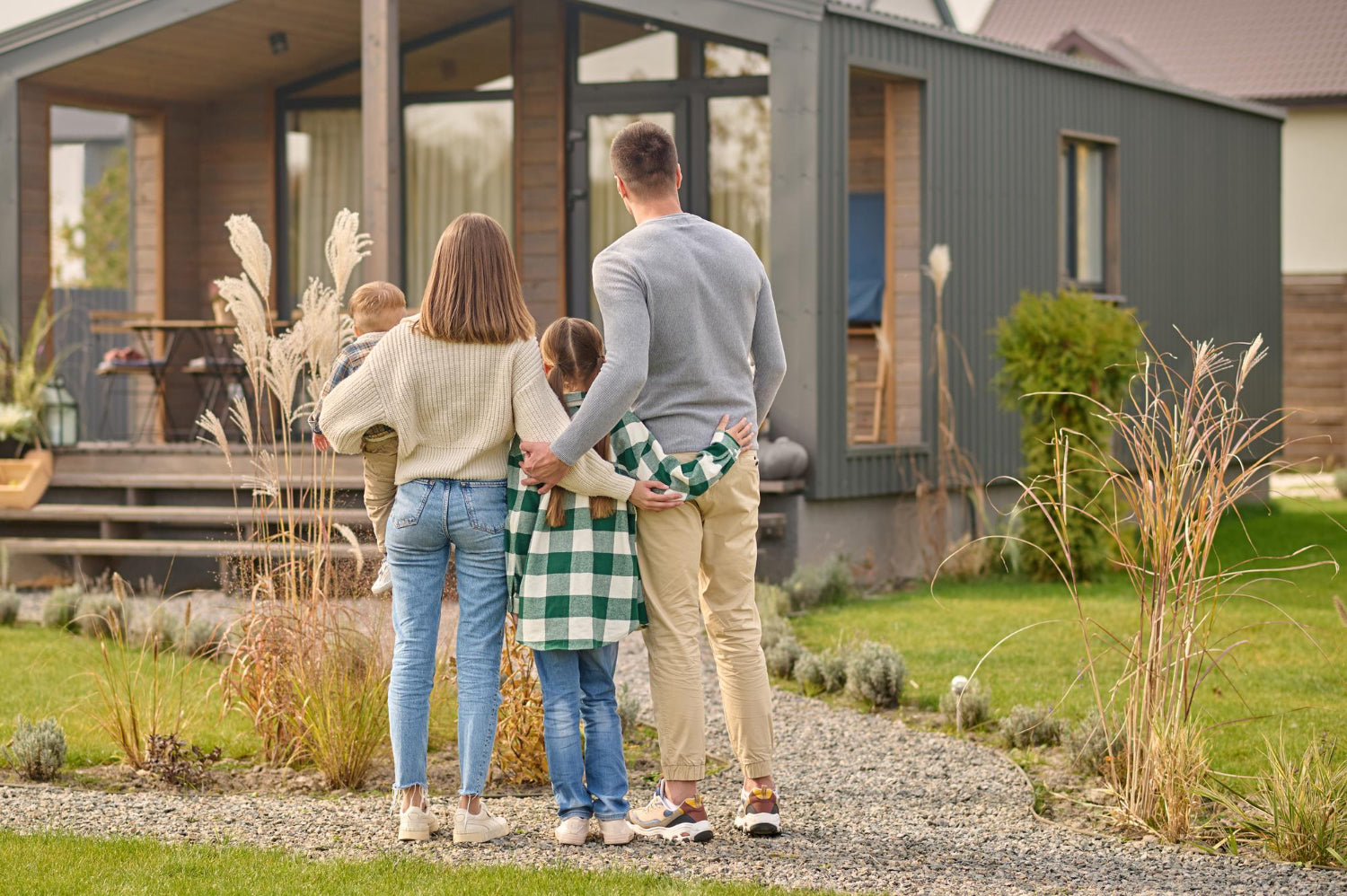 Family of four standing on a gravel path, looking at their modern, dark grey modular home and backyard.